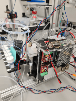 Photo of the automated liquid exchange system, a compact electronics module with circuit boards and control wiring mounted alongside tubing and a multi-well plate on the microscope stage.
