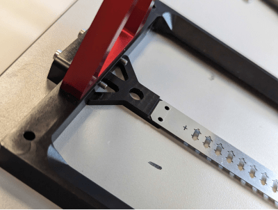 Photo of a metal sample-holding bracket with mounting slots, positioned under a red clamp arm on the light-sheet microscope stage.