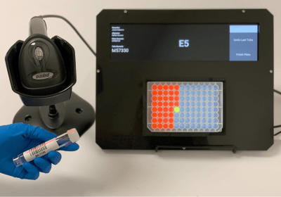 Photo of the Well-Lit device displaying a 96-well plate grid with one illuminated well, alongside a barcode scanner and a gloved hand holding a sample tube.