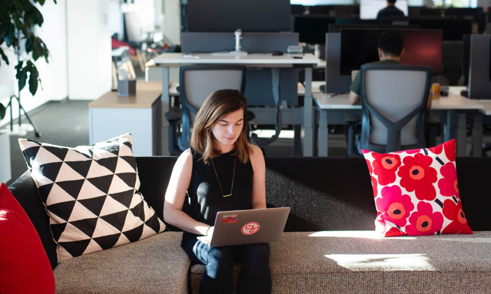 A woman works at a laptop on a couch in an office enviroment.