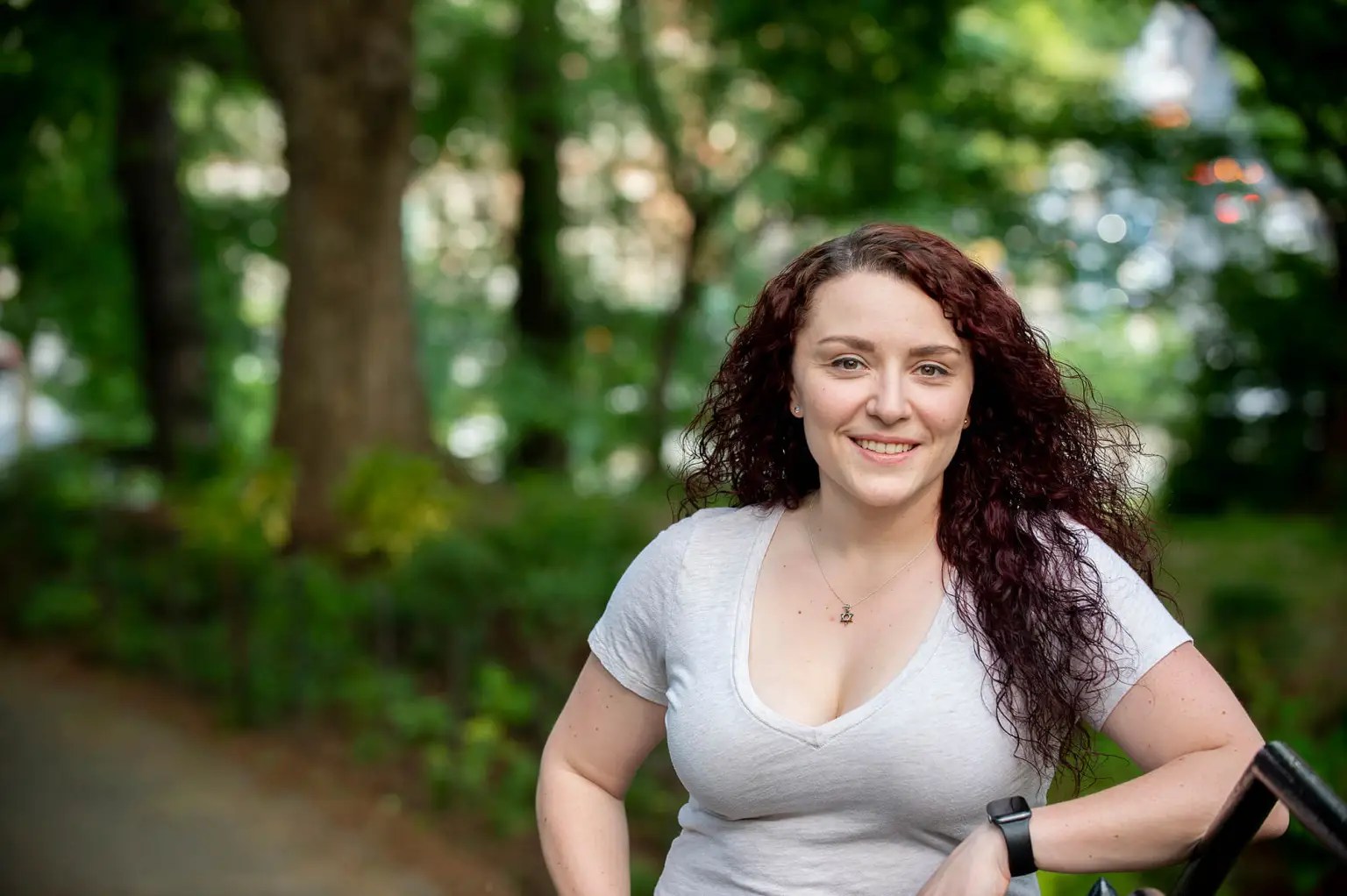 A woman with red-brown hair and in a white shirt smiles in a wooded area.
