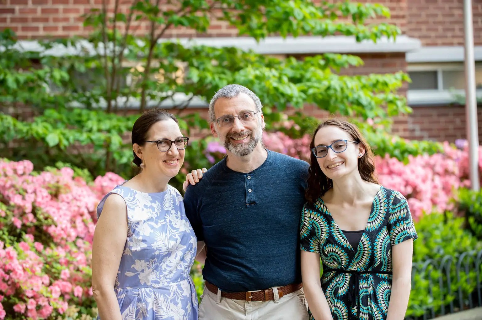 A family of three smile for a photo with greenery and pink flowers in the blurred background.
