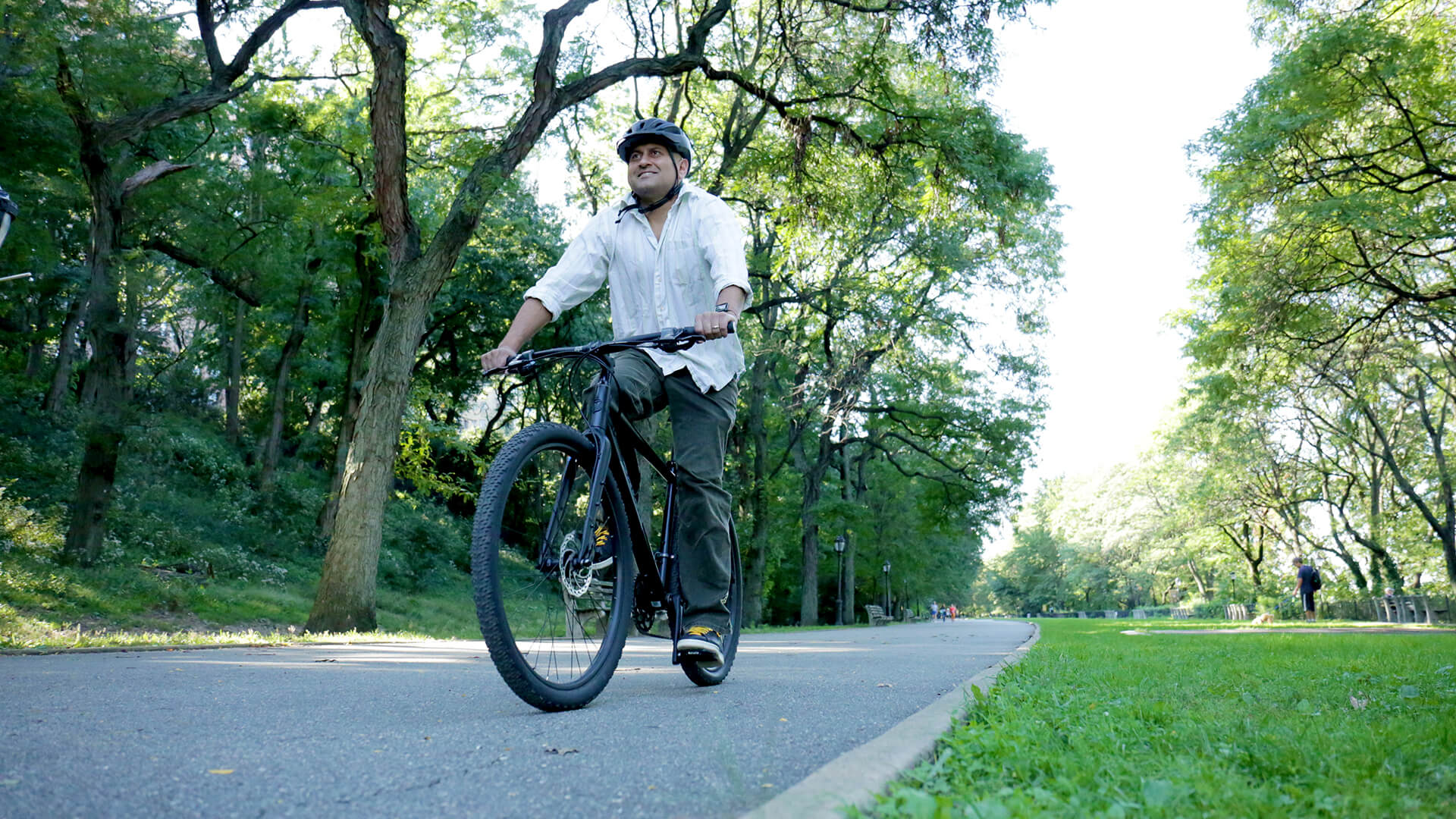 A man rides a bike wearing a helmet. Green trees are in the background.