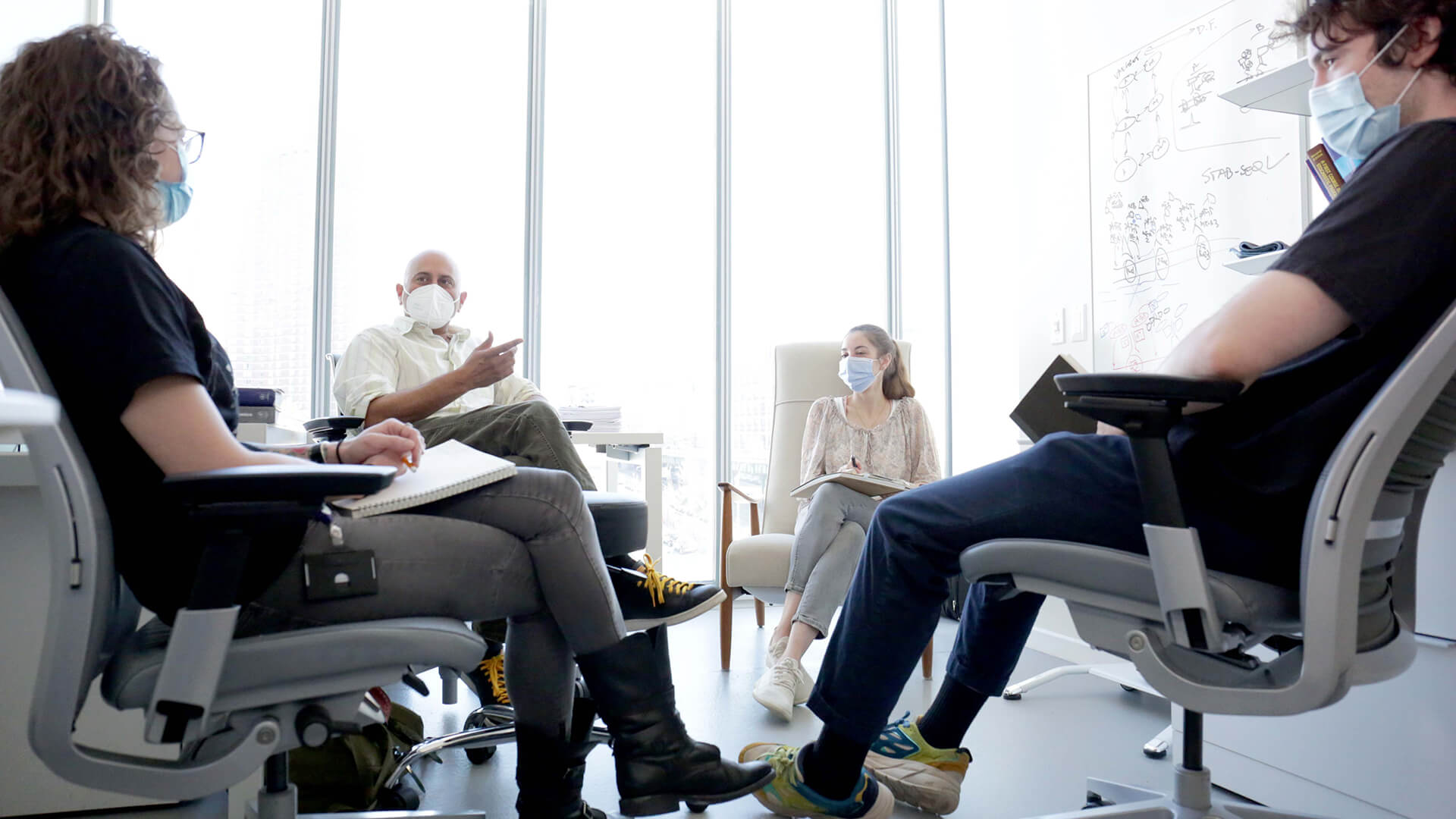 A group of people wearing masks sit in a circle in an office. 