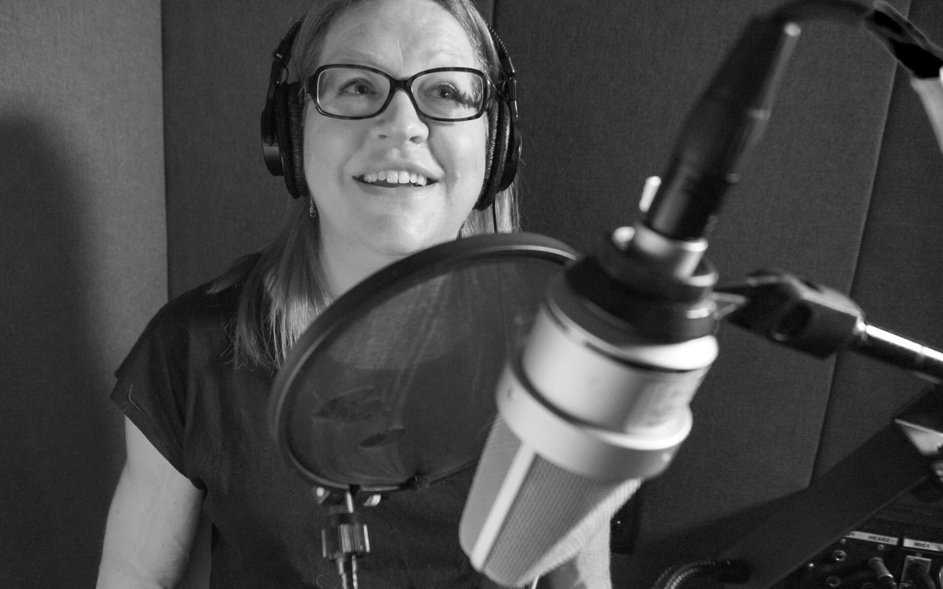 A black and white photo of a woman wearing glasses that’s sitting in front of a microphone and pop filter. 