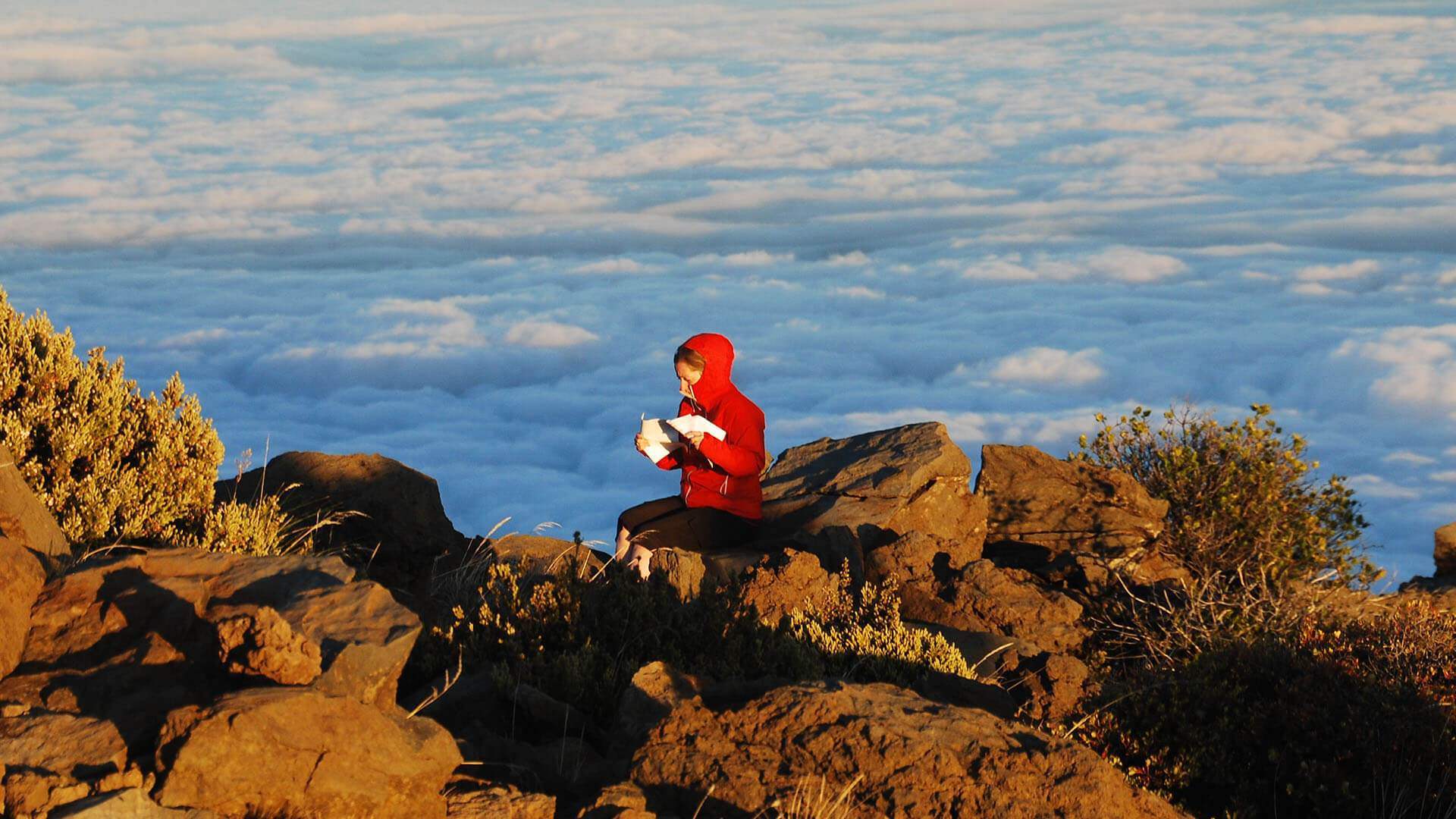A woman in a red jacket sits on a rock with clouds in the background.