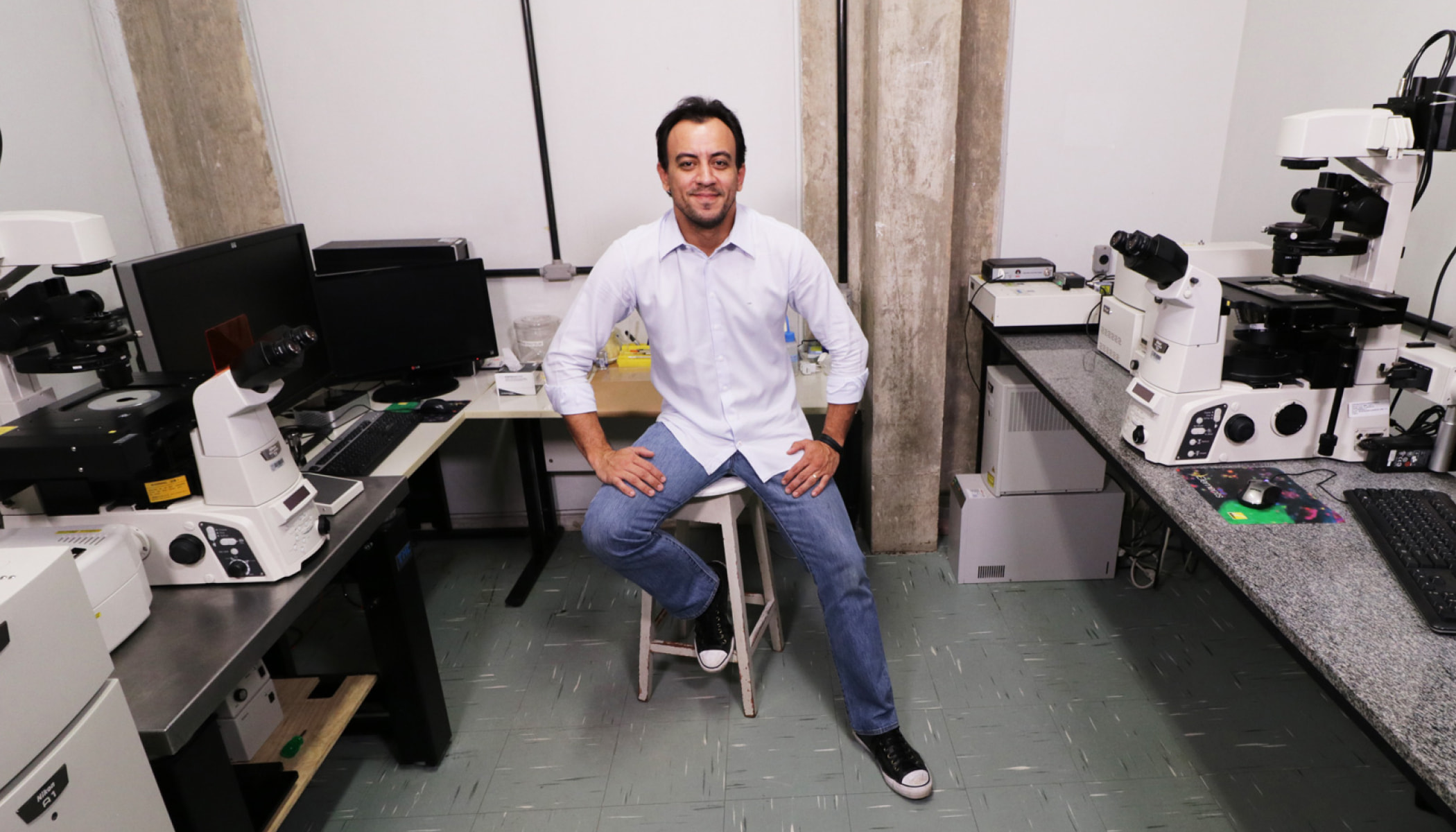 Biologist Gustavo B. Menezes of Brazil sits on a stool in a lab, with microscopes and computer screens.