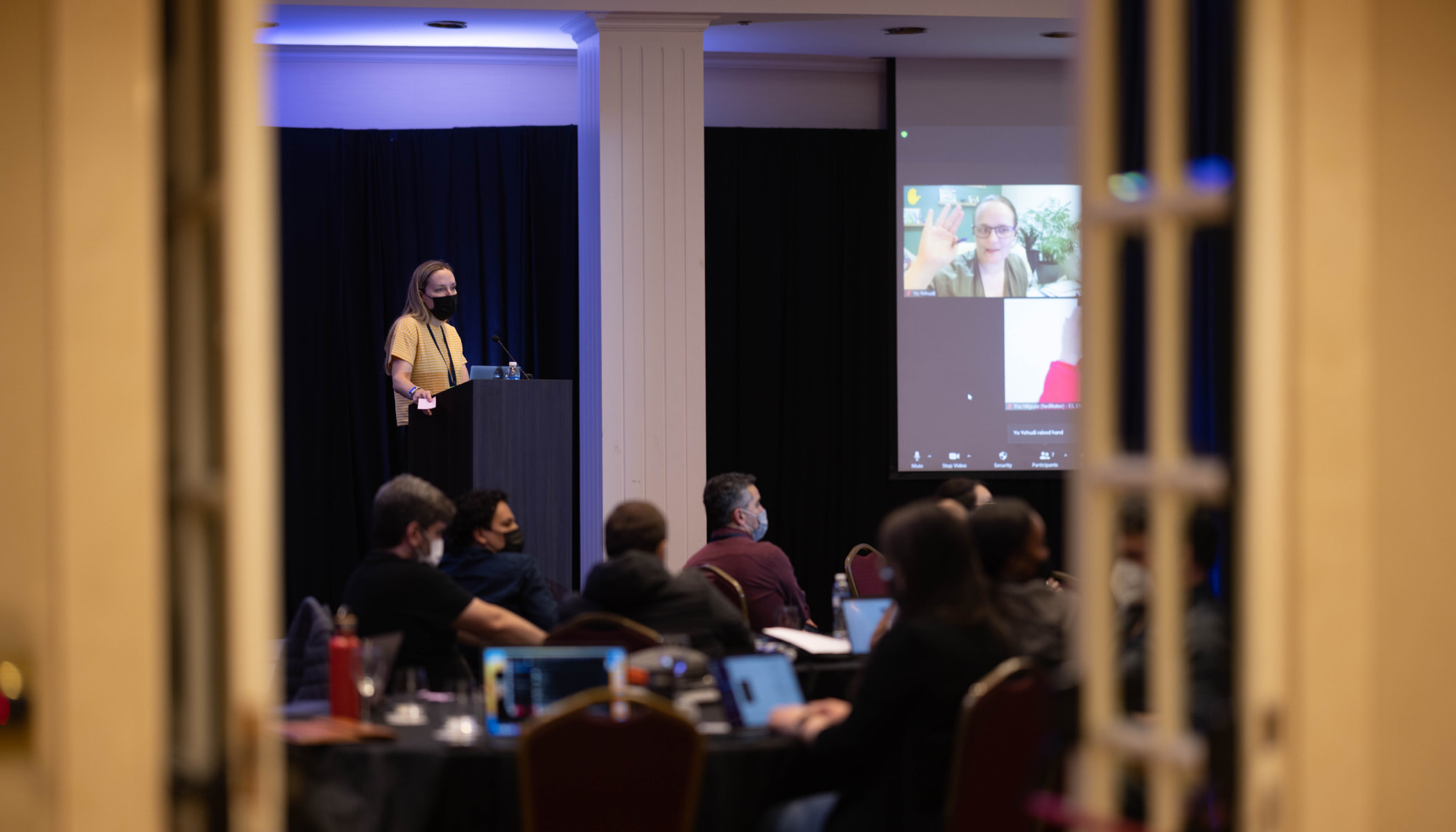 A shot through an open doorway of a speaker at a podium and a room full of viewers. There’s a partial view of a large screen with Zoom call participants.