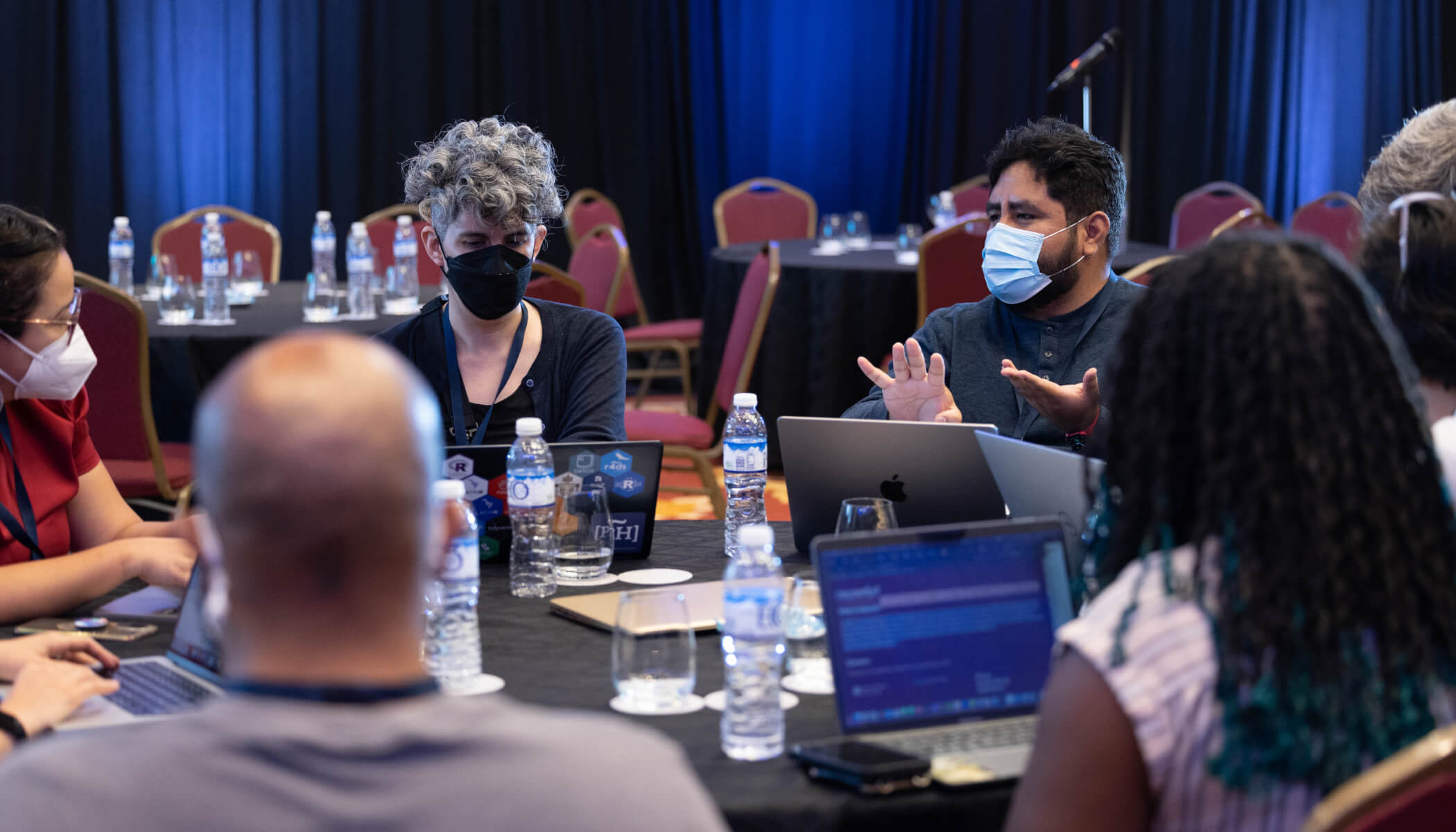 Event attendees wearing masks sit in discussion around a table with their laptops.