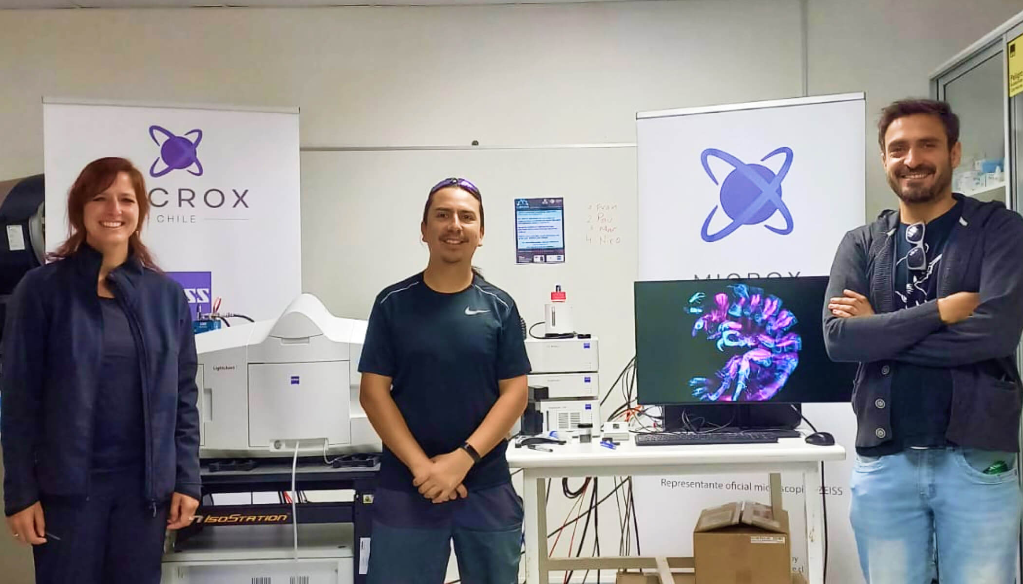 Alenka Lovy and colleagues smile and stand in front of a light-sheet microscope with a computer screen showing live imaging of a developing organism in bright purples and blues.