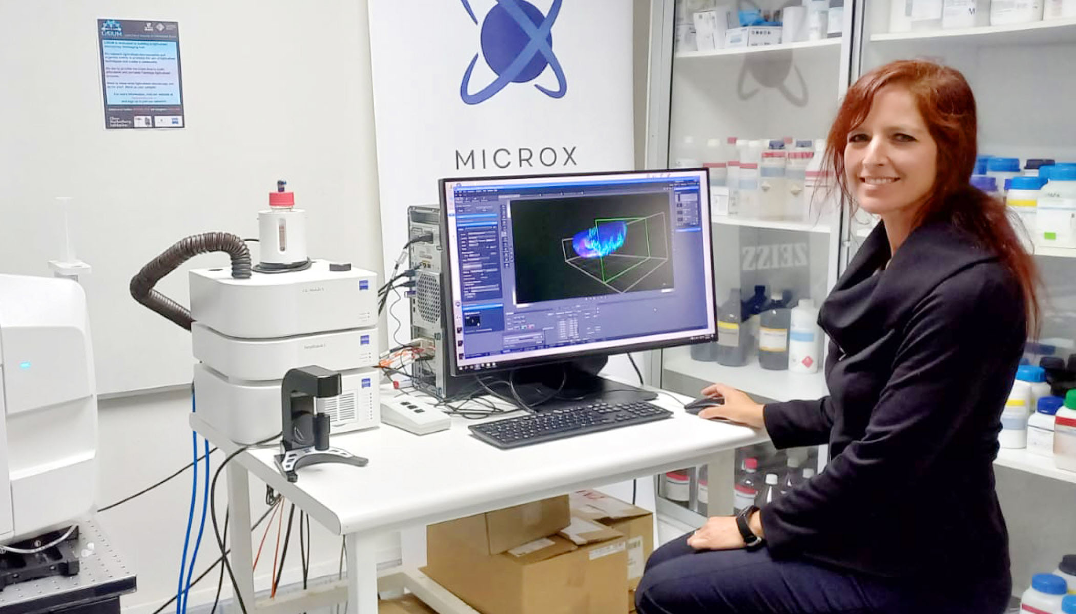 Alenka Lovy smiling in a lab, posing in front of a computer with imaging software open next to a light-sheet microscope.