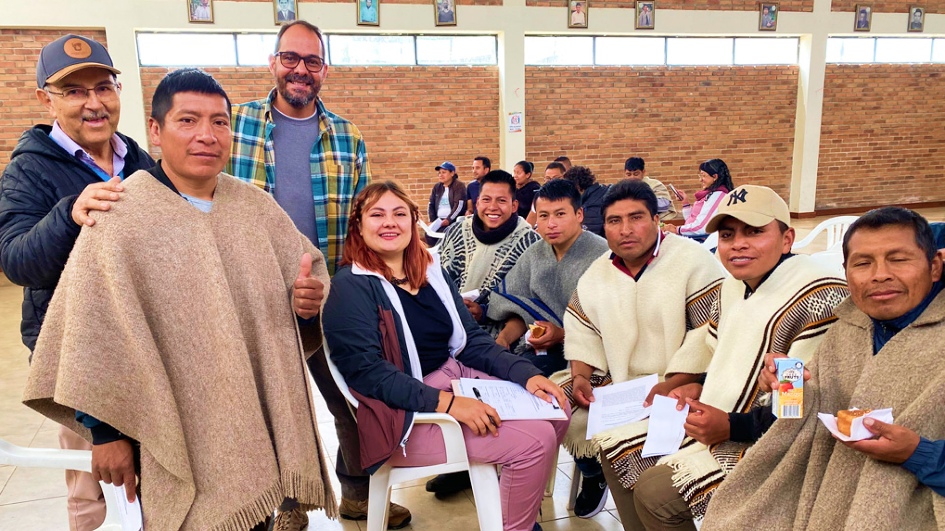 Alt-text: Andrés Moreno-Estrada and his research team sit and stand with members of the Pastos community in the Andes, smiling and holding papers during a meeting indoors.
