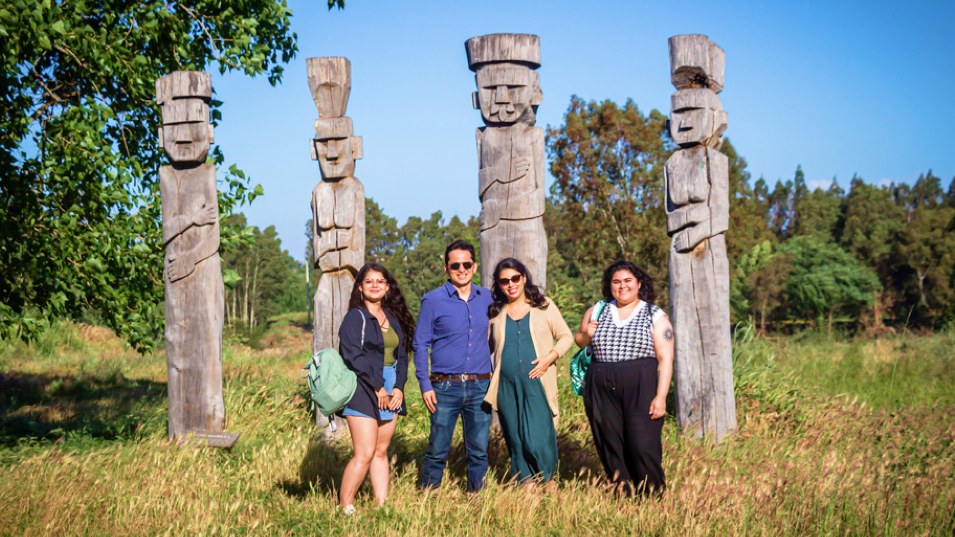 Four members of a Chilean research team stand in front of five tall wooden statues in a grassy clearing, surrounded by trees and blue sky.