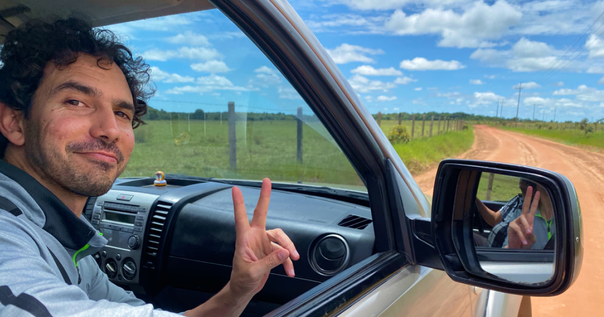 Carlos smirking out of the passenger side window of a silver car, holding his hands up in a peace sign