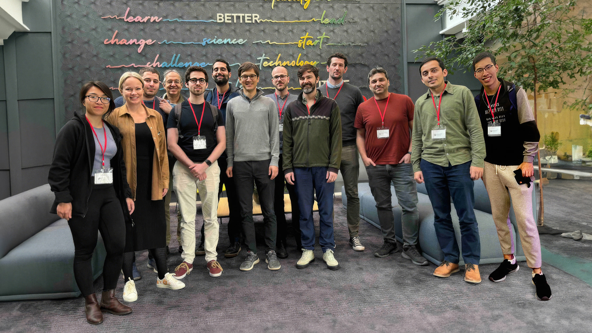 A group of individuals standing together in an office. Most wear red lanyards and an art installation is in the background.