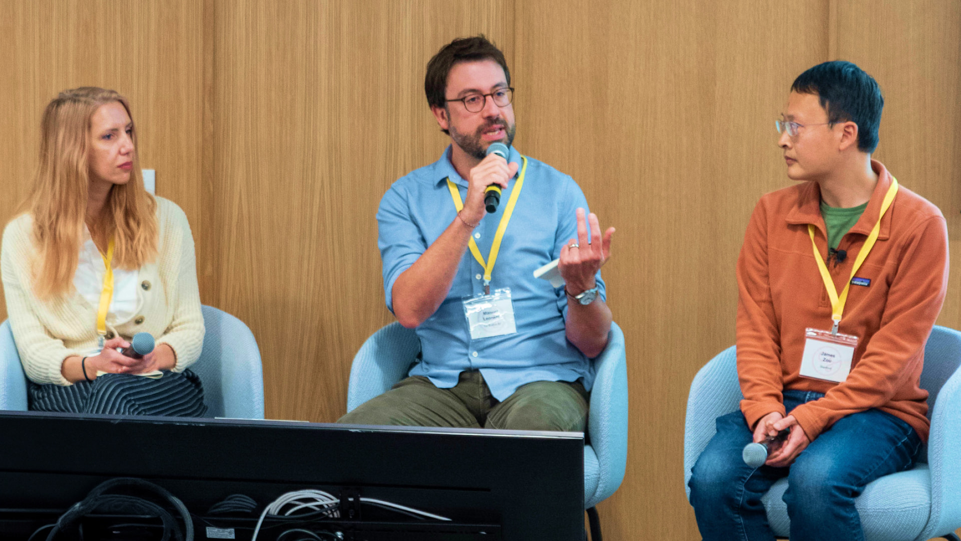 Three people are seated on a panel, engaged in a discussion; a man in the middle speaks into a microphone while gesturing, with a woman on the left and a man on the right listening attentively.