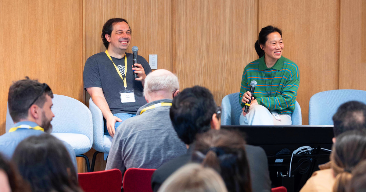 Two people are seated on a panel, both holding microphones and smiling; the man on the left, Boris Power, is wearing a T-shirt with a logo and a lanyard, while the woman on the right, Priscilla Chan, is wearing a green striped shirt. An audience is visible in the foreground.