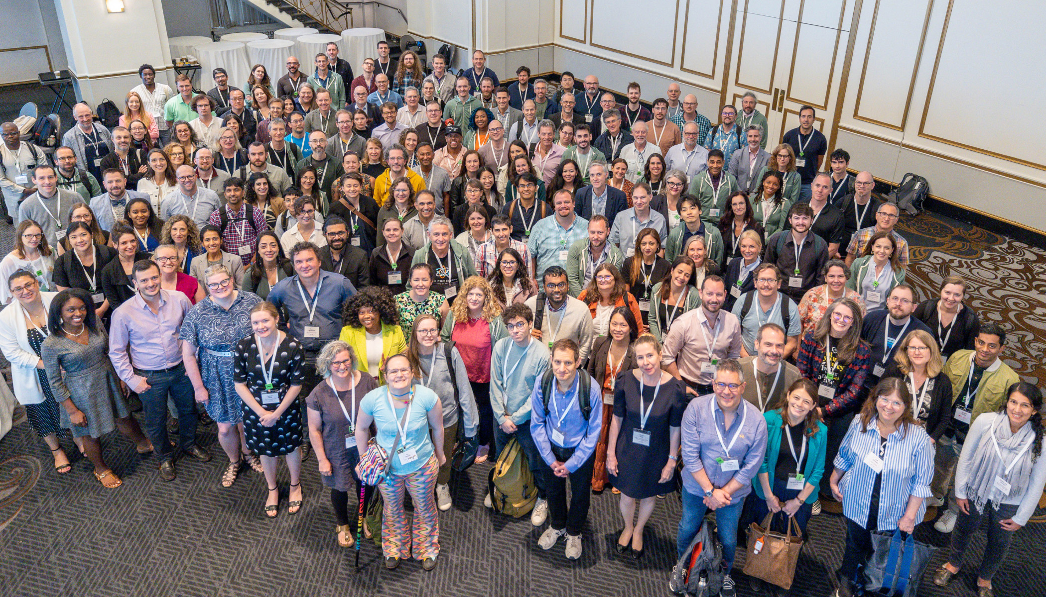 A large, diverse group of people, all wearing conference badges, are gathered together in a ballroom, smiling and looking up at the camera for a group photo.