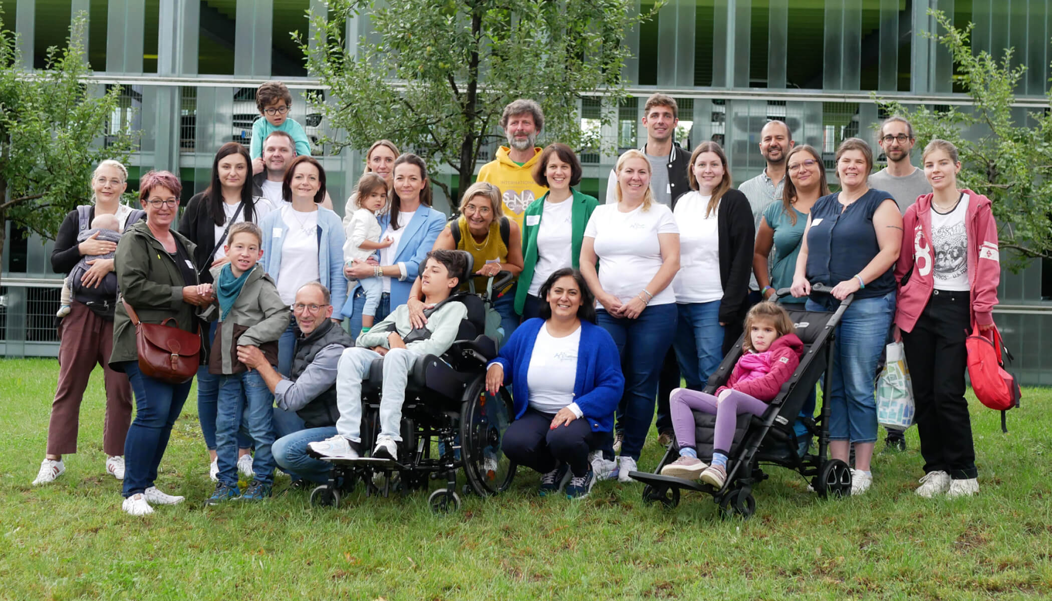 A group of families, including children and adults, pose outdoors. The background shows a modern building with trees in the foreground.