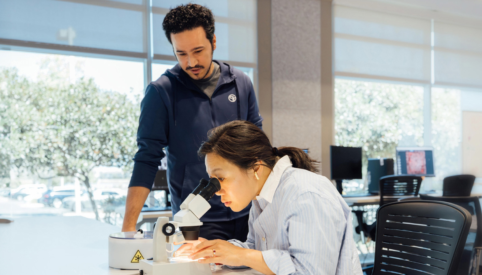 Priscilla Chan looks into a microscope while a colleague observes her in a well-lit laboratory at the CZ Imaging Institute.