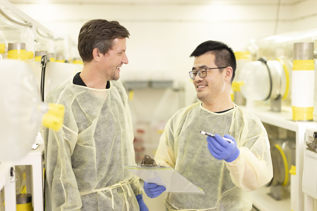 Scientist Noah Palm stands next to a lab member dressed in yellow PPE gowns and blue gloves. They smile at each other. A lab is seen in the background.