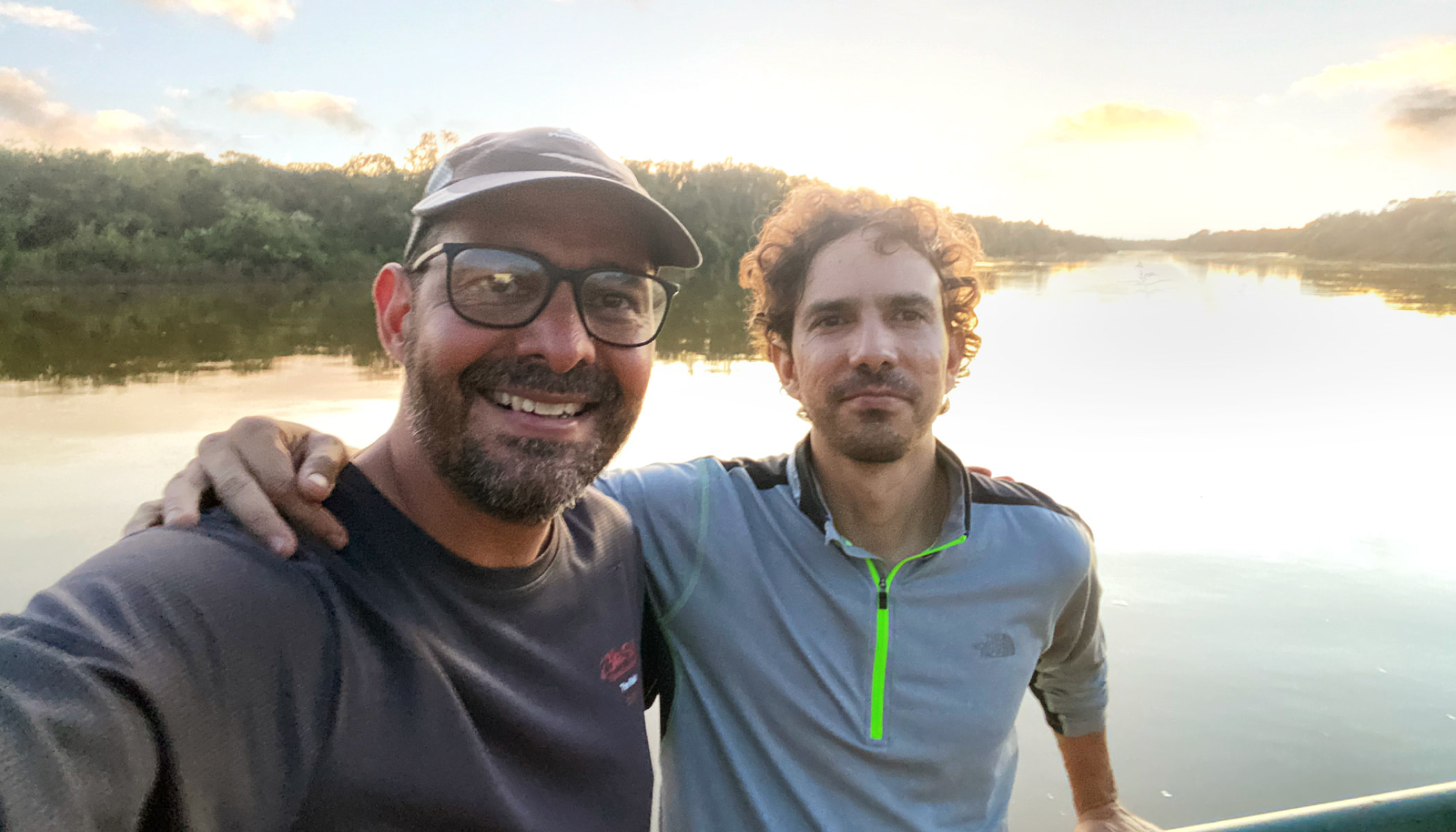 Selfie in front of a sun-reflecting river with Andrés wearing a cap and glasses smiling next to Carlos, who is leaning on the edge of the boat with an arm around Andrés.