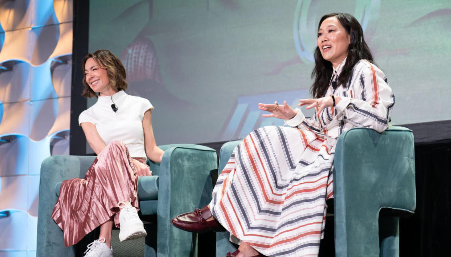 Priscilla Chan and Emily Chang speak on stage at SXSW. Priscilla gestures while talking, wearing a striped dress, and Emily smiles beside her, wearing a white top and pink pleated skirt.