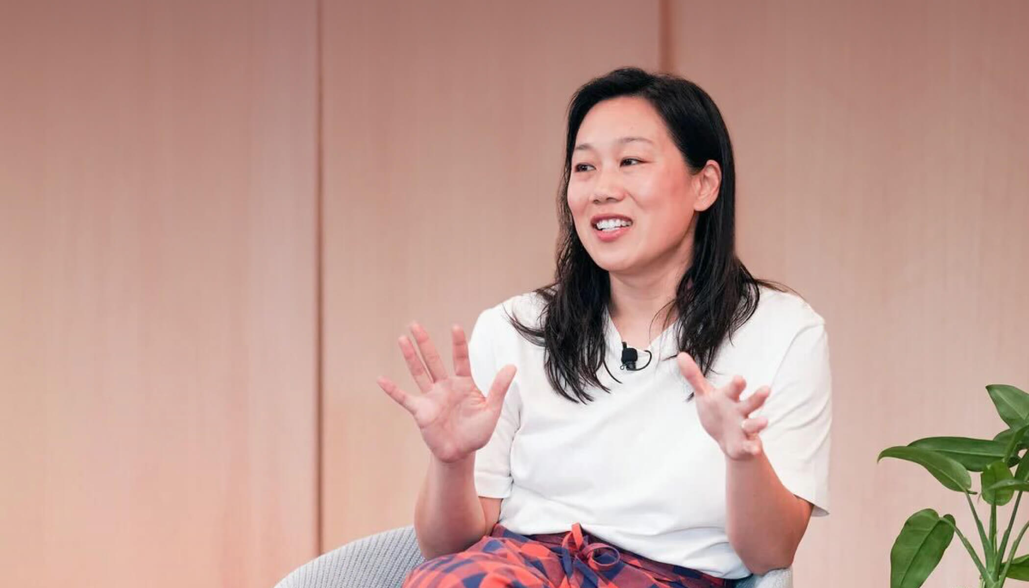 Priscilla Chan sits in a chair and gestures with her hands. She wears a black and red checkered skirt and a white T-shirt.