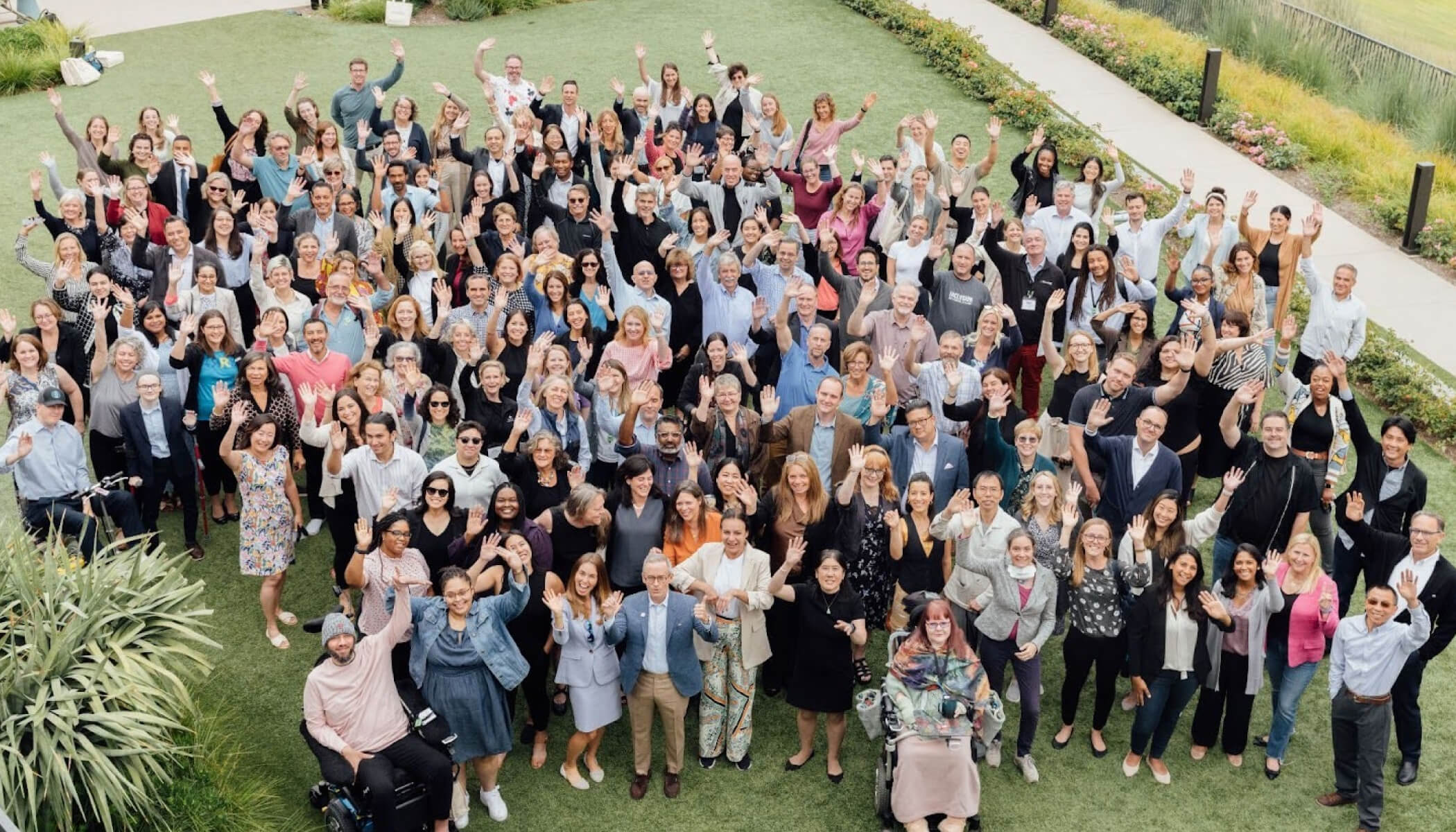 A large group of people stand together outside on grass, smiling and waving at the camera in a celebratory pose.