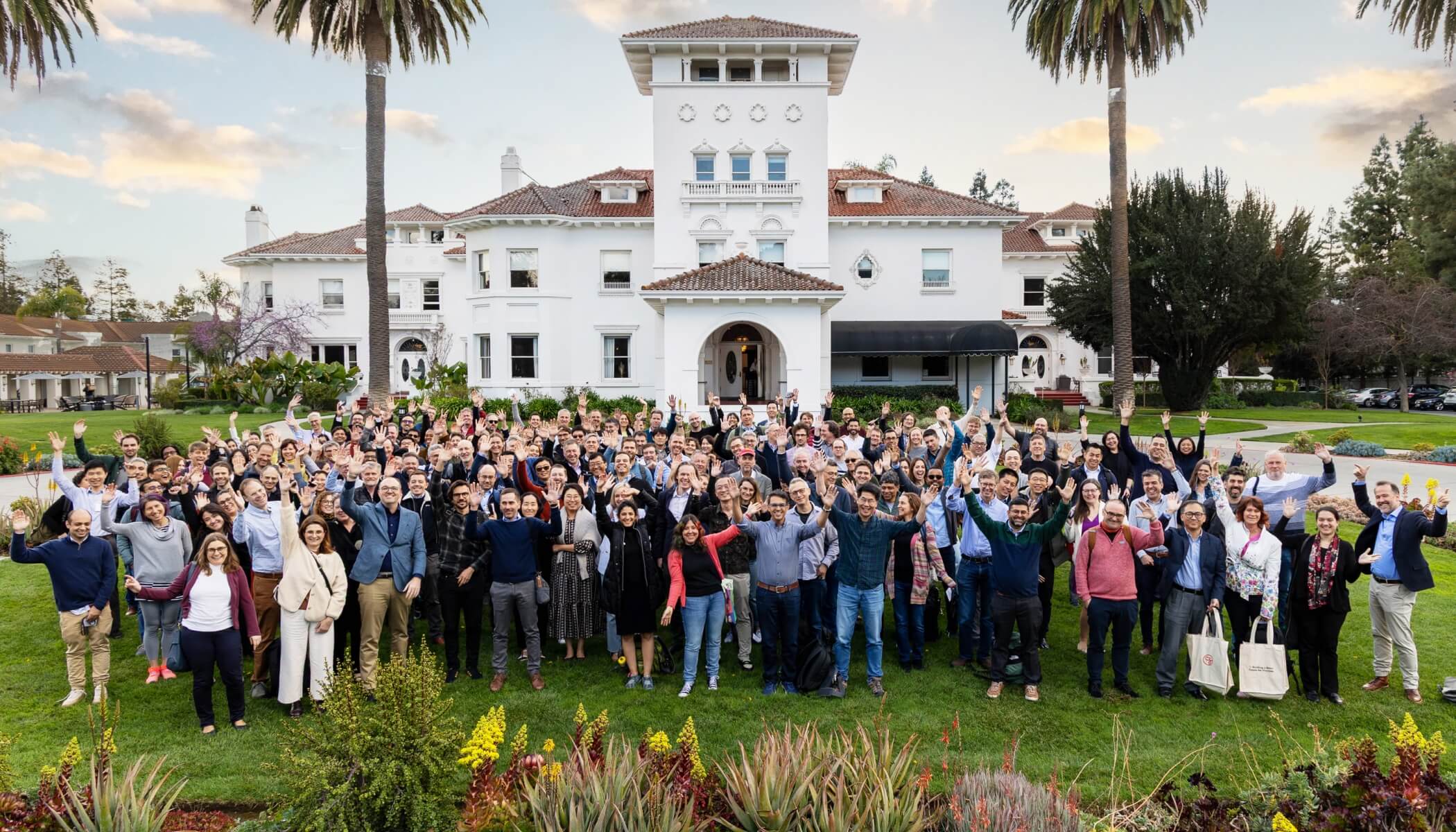 A large group of people stands outdoors under tall trees, smiling and waving at the camera.