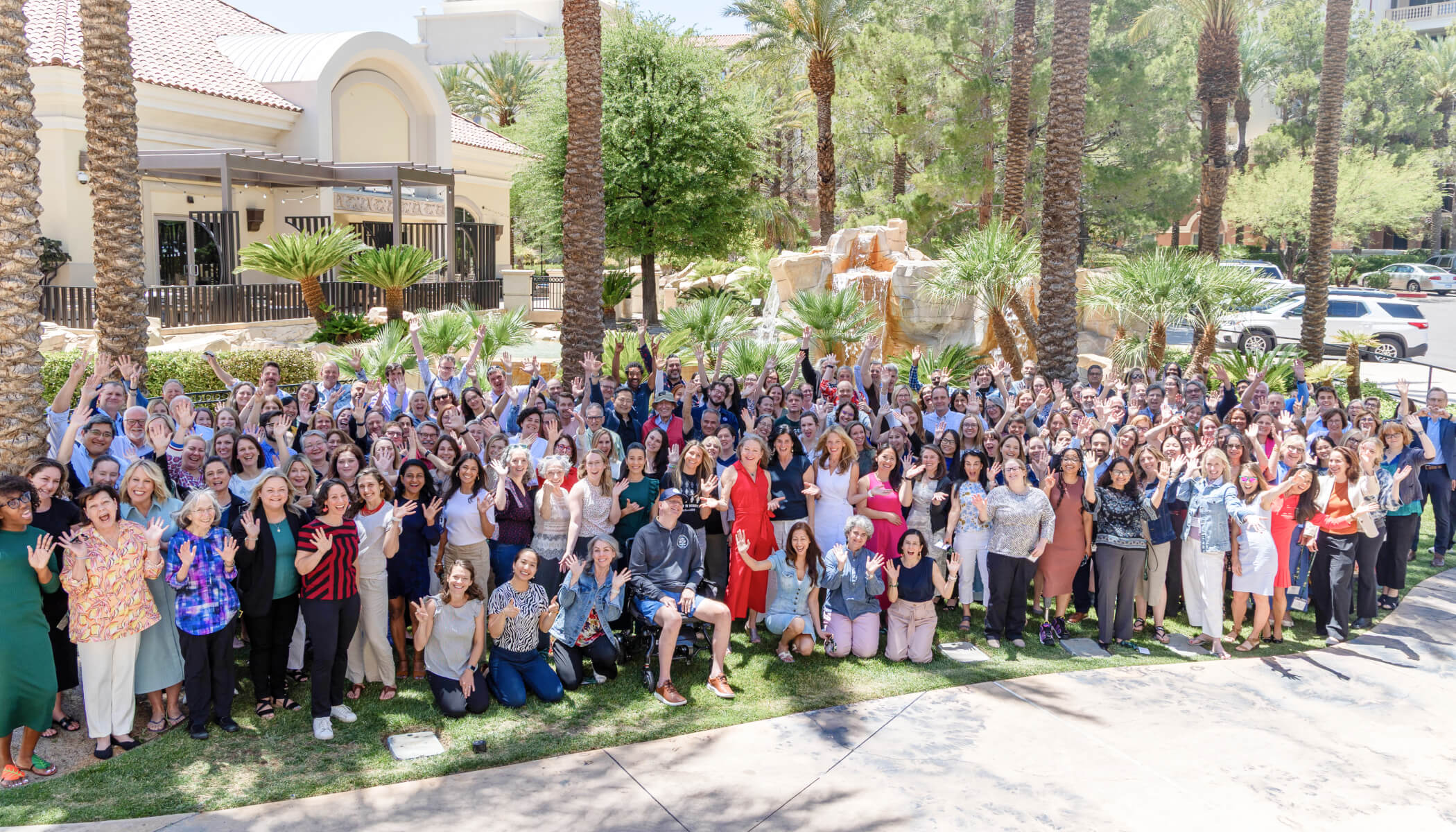 A large group photo of people standing together, smiling and waving outdoors among palm trees.