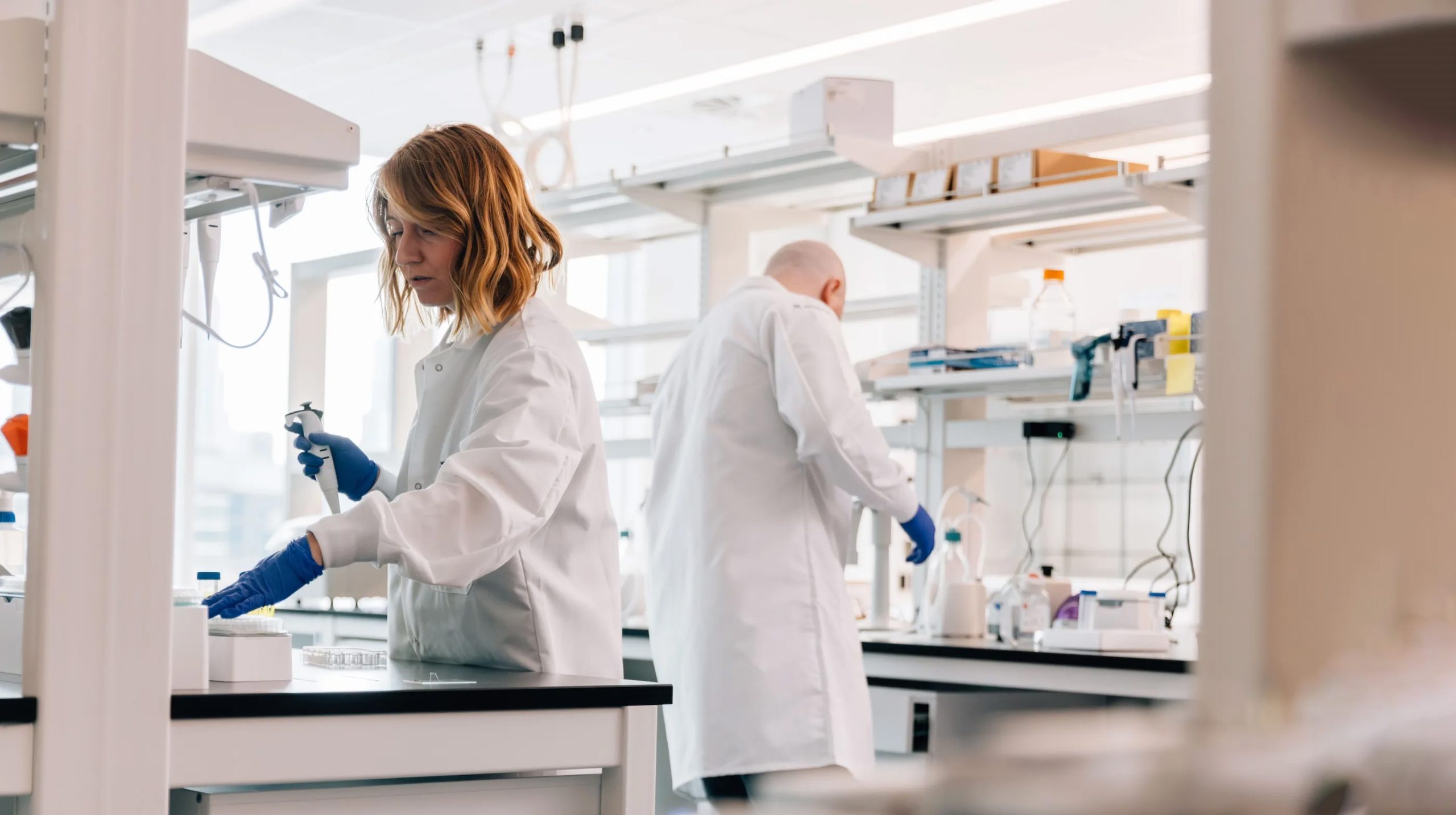 Scientists in a laboratory wearing white coats and gloves perform pipetting and sample preparation at a bench with standard molecular biology equipment.