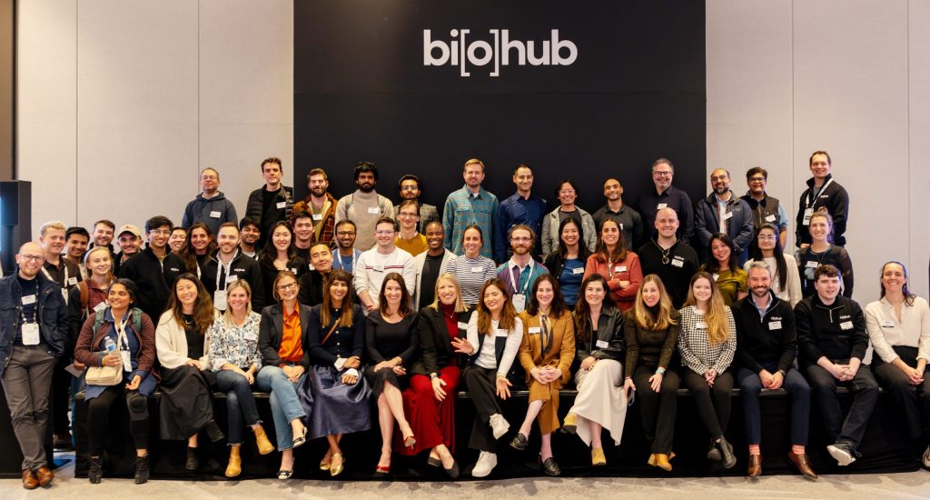 Large group photo of the Biohub team, arranged in three rows in front of a wall with the Biohub logo